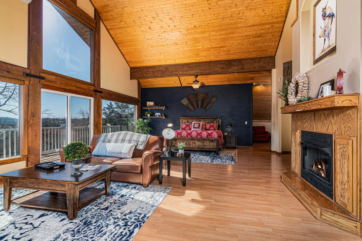 A spacious, elegantly furnished living room featuring a fireplace, large windows with scenic views, wooden beams, and stylish furniture. The room exudes comfort and warmth, inviting relaxation by the fire or enjoying the view.