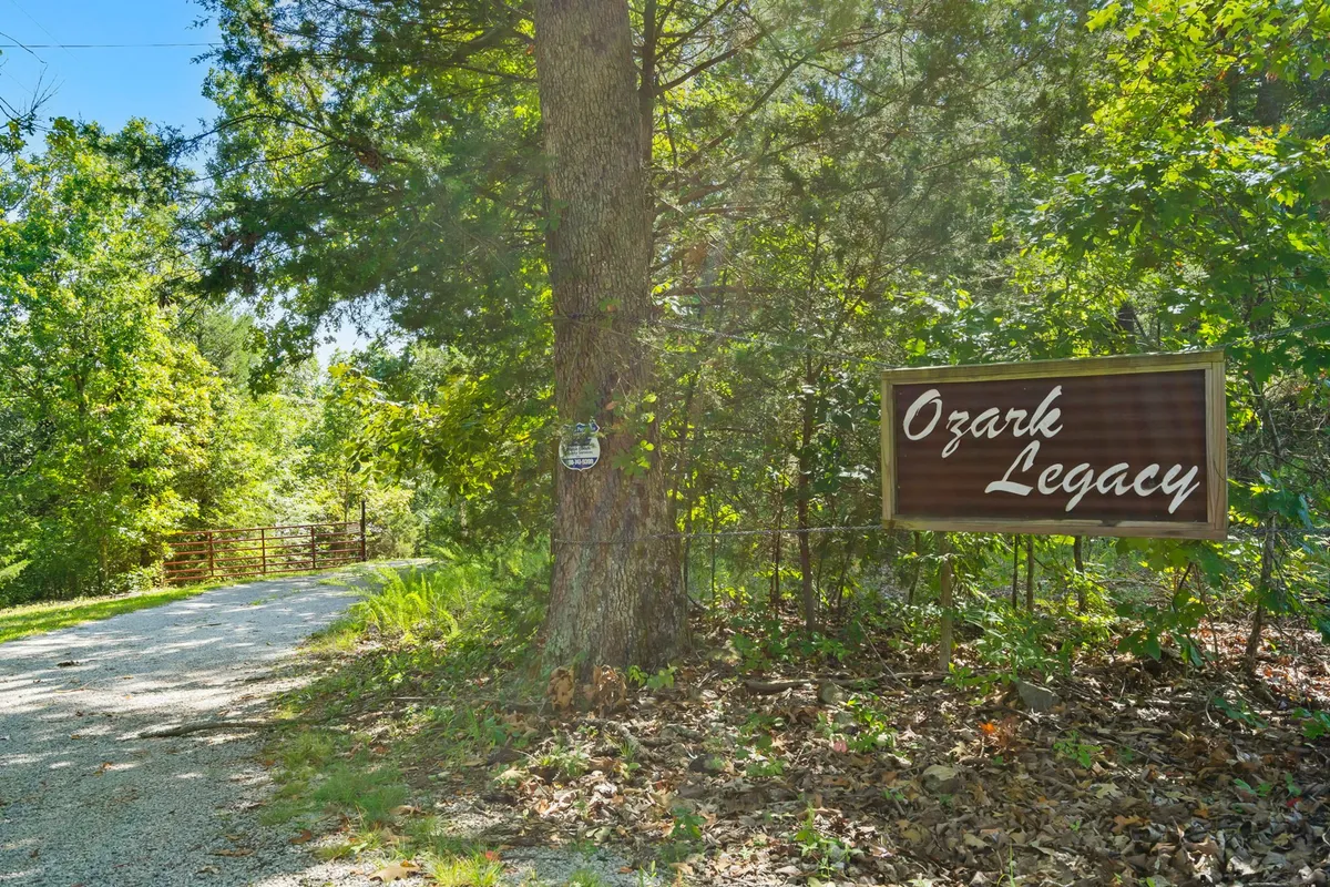 An exterior view featuring the Ozark Legacy sign at the end of a gravel driveway, surrounded by mature trees. The setting feels secluded and tranquil, evoking a sense of arrival and the promise of exploration in natural surroundings.