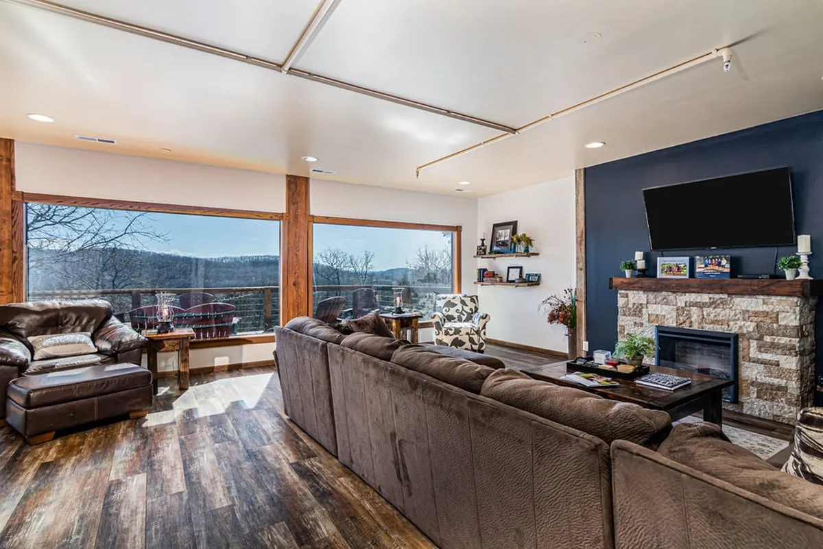 A cozy living room featuring a comfortable seating area, large windows with panoramic outdoor views, a stone fireplace, and decorative shelving. The space is perfect for relaxing, entertaining, and enjoying sunset vistas with loved ones.