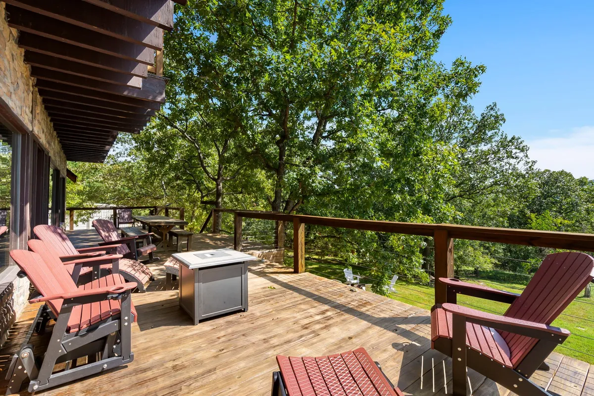 A calm, relaxed exterior setting with a wooden deck, red Adirondack chairs, an outdoor table, and green trees in the background. The scene evokes moments of peaceful relaxation or casual socializing in the fresh air.