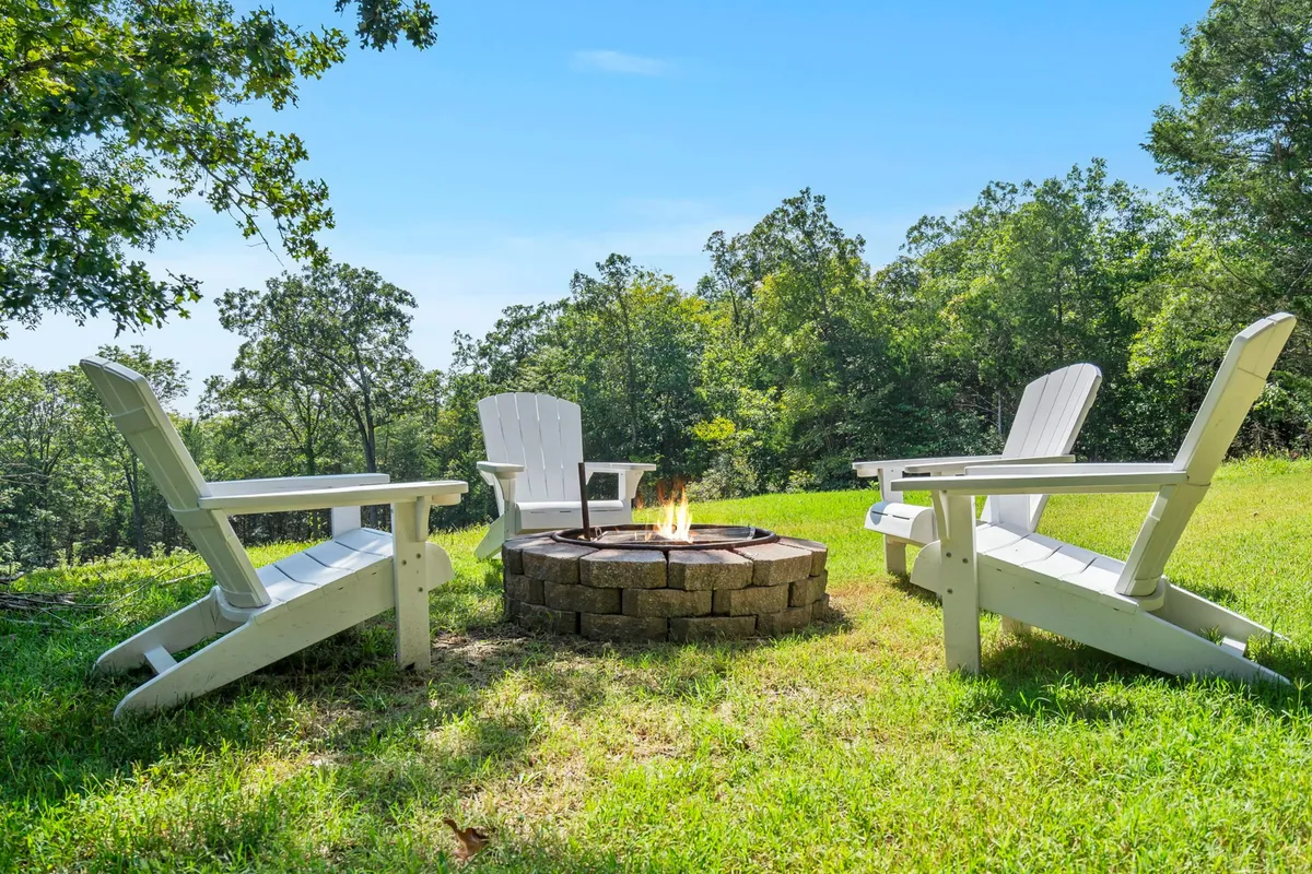 A cozy fire pit ringed by Adirondack chairs sits in a green, wooded clearing, ready for campfire gatherings, storytelling, and marshmallow roasting as dusk falls.