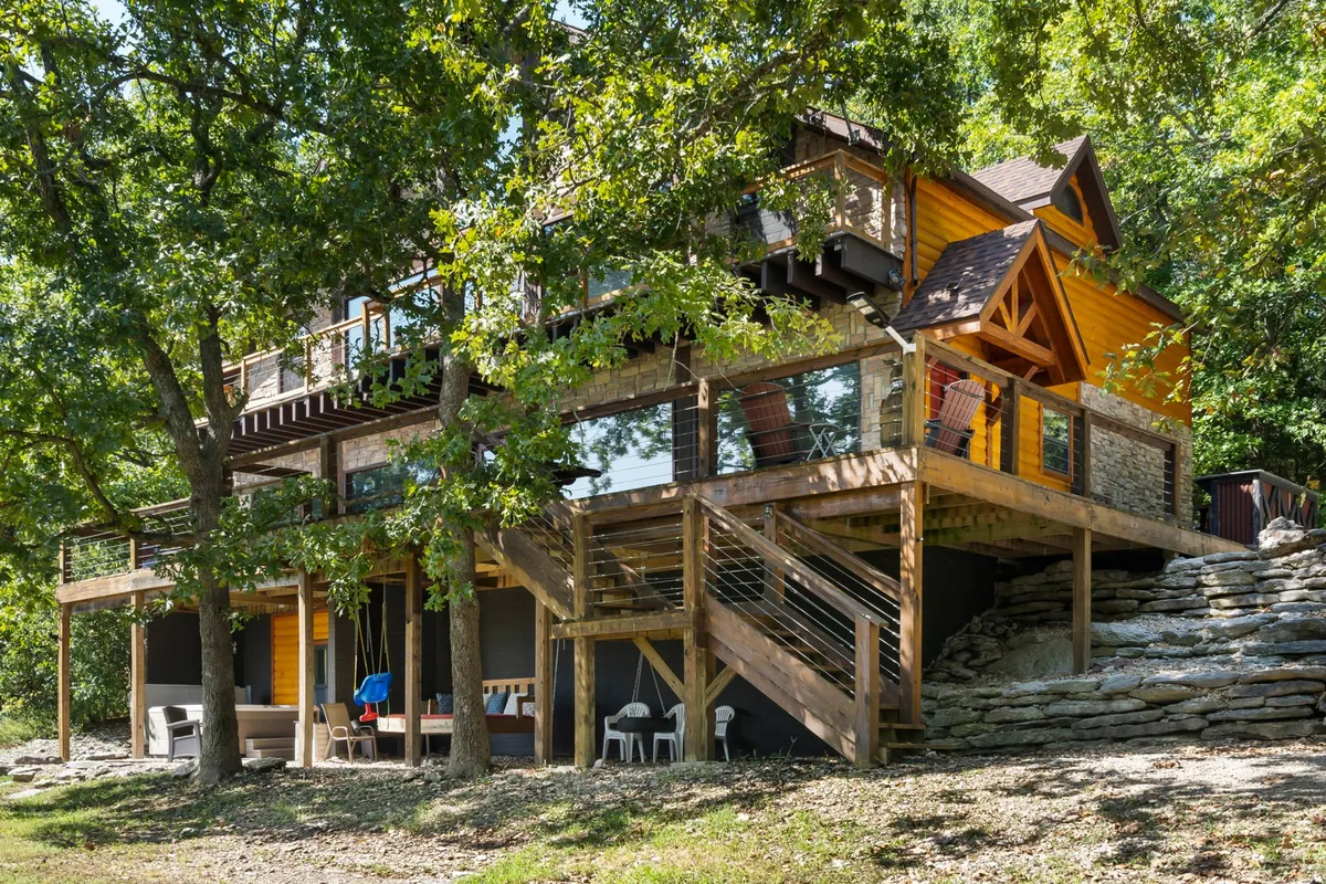 A welcoming exterior view showing the home's wooden deck, large windows, colorful facade, and lush surrounding trees. The deck is set up for outdoor dining and relaxation, offering a glimpse of the inviting spaces inside, as well as the beautiful natural environment all around.