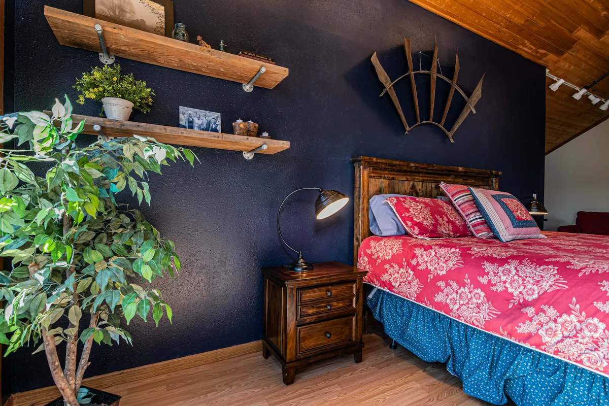 A welcoming bedroom with a bed dressed in floral bedding, wooden shelves holding decor and books, decorative wall accents, a nightstand with a lamp, and a potted plant. The setting is cozy and homey, ideal for relaxation and quiet moments.