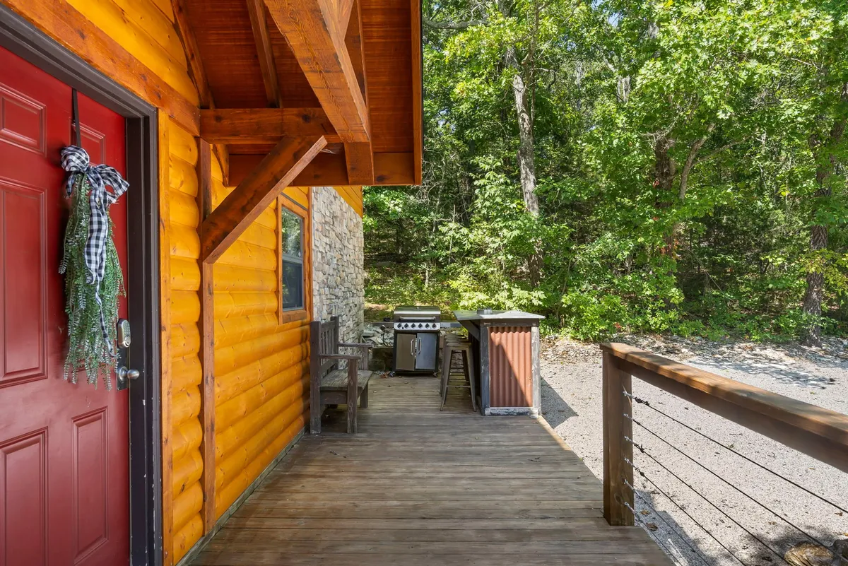 This image features the exterior of the property with a prominent red front door, a spacious wooden deck equipped with a grill, and an inviting setting surrounded by trees. The deck provides a casual, social outdoor space ideal for group barbecues and gatherings after attending events or exploring local attractions.